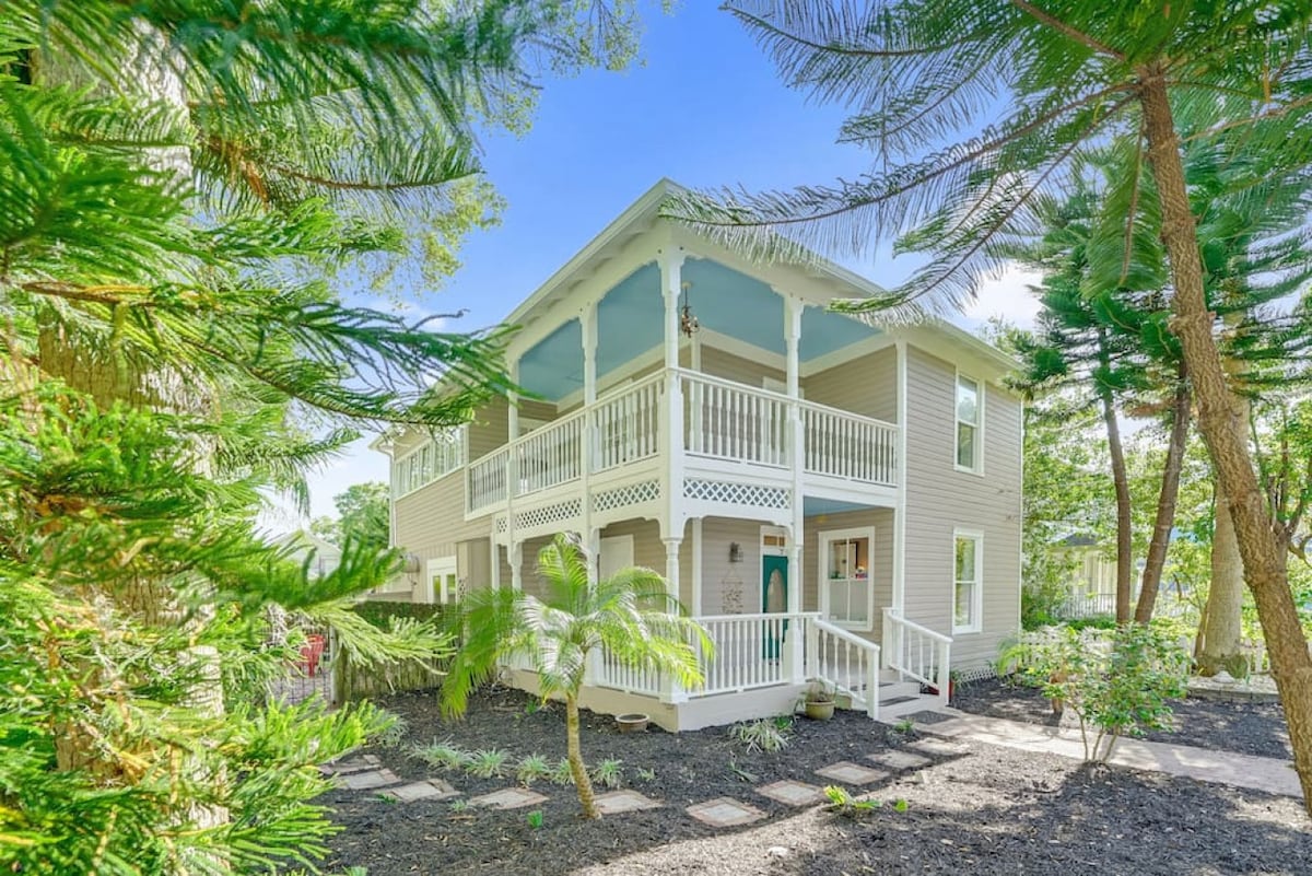 The exterior of a two-story Victorian home is framed by lush greenery, including palm trees and shrubbery. The home features a covered porch with white railings and a blue ceiling, inviting guests to relax while enjoying the serene surroundings.