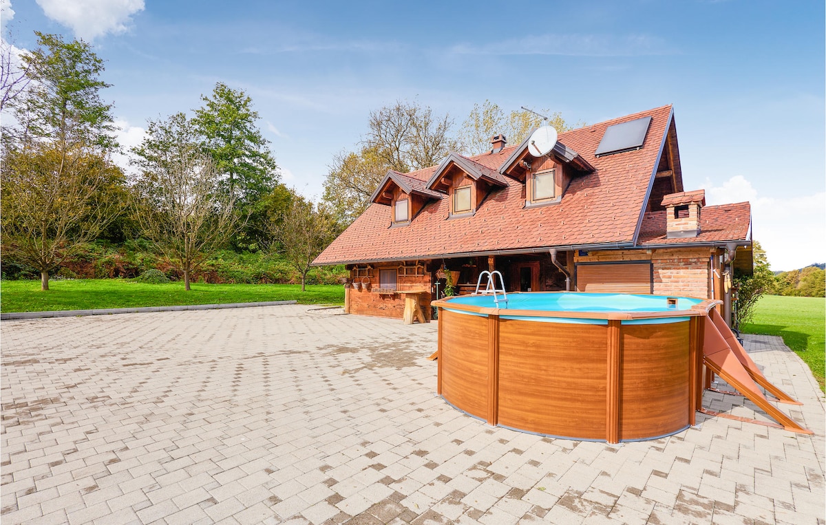 A warm-toned, rustic home is featured beside a circular above-ground pool. The pool area is surrounded by a paved patio and grassy landscape, with trees and greenery in the background. The house displays a combination of wooden accents and a red tiled roof.