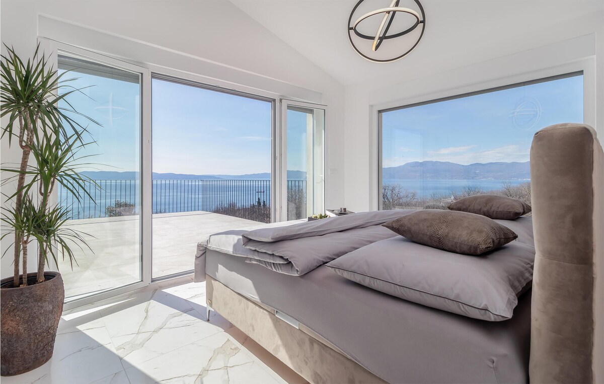 A modern bedroom features a spacious bed with soft bedding, framed by large glass doors offering expansive views of the sea. A potted plant adds a touch of greenery, and the floor displays elegant marble tiles, complementing the serene atmosphere.
