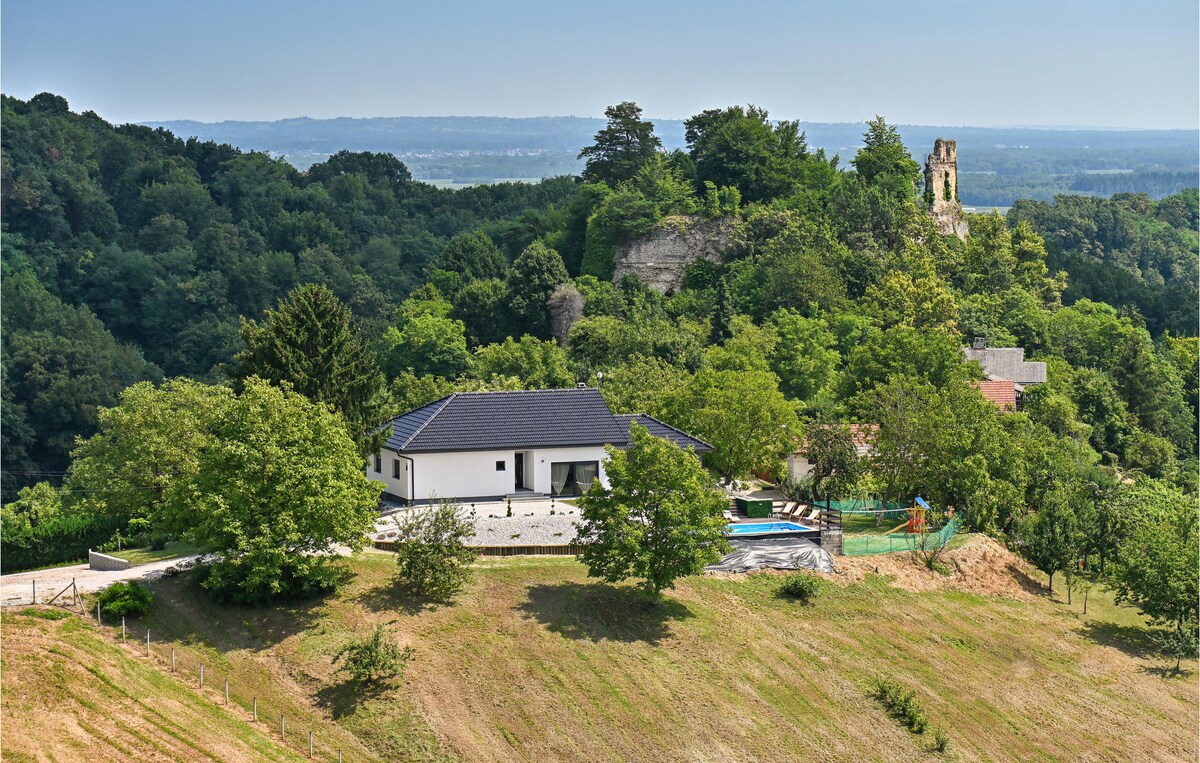 The holiday home is set amidst greenery, with trees surrounding the property. A private pool can be seen in the outdoor area, and a historic castle tower is visible in the background, adding charm to the scenic countryside landscape.