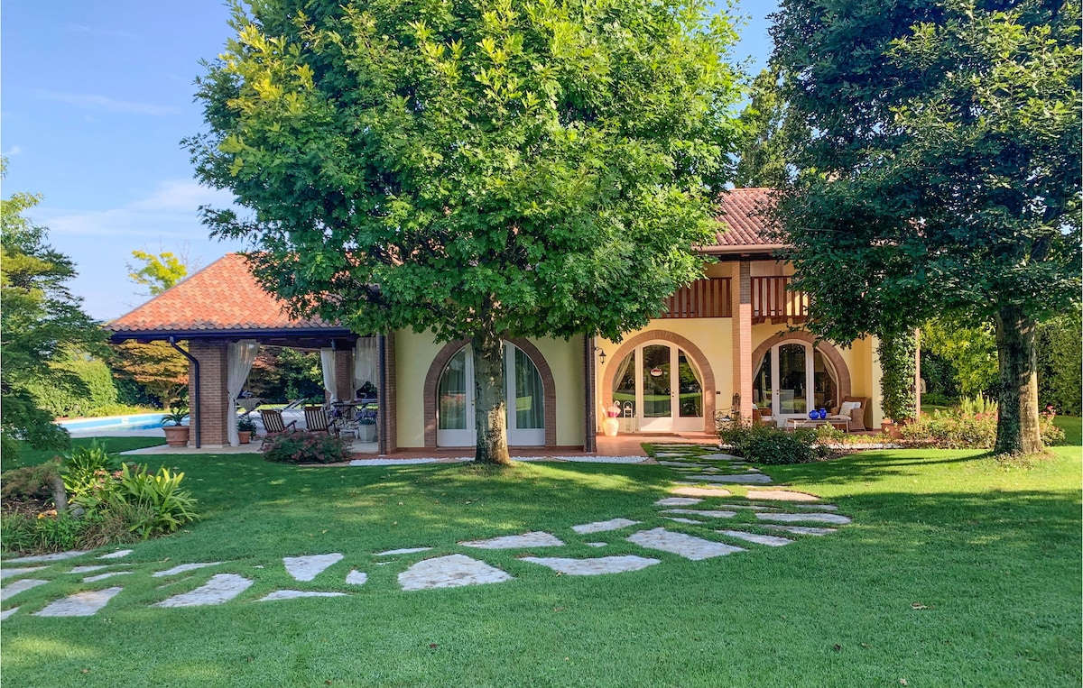 The cottage is framed by lush greenery and two large trees, showcasing a harmonious blend of architecture and nature. A well-maintained lawn leads to the inviting entrance and outdoor seating areas. The swimming pool is visible in the background, enhancing the tranquil setting.