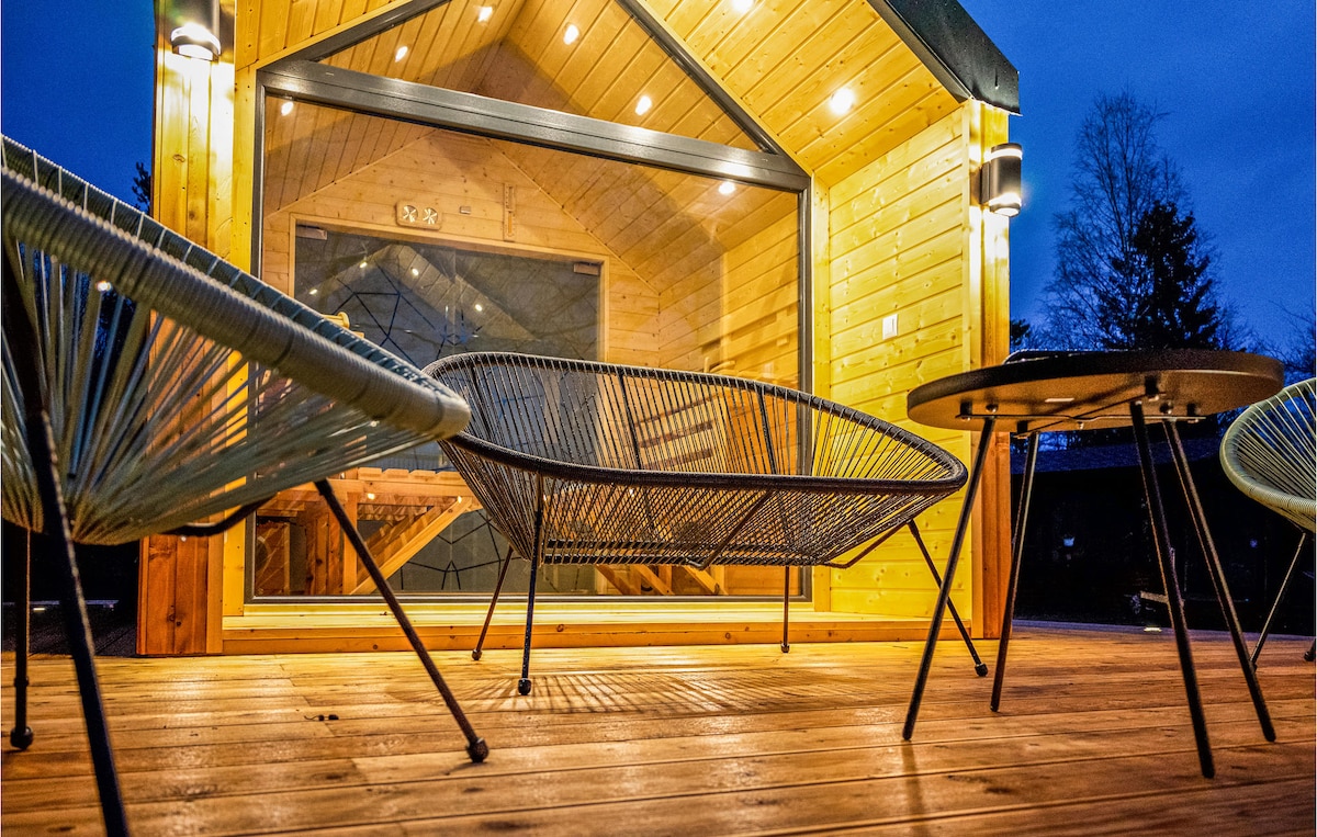 A warmly lit outdoor area features several contemporary lounge chairs arranged around a modest table. The cabin structure is visible in the background, showcasing large windows that enhance the inviting atmosphere. Soft lighting highlights the space, creating a cozy setting for gatherings.