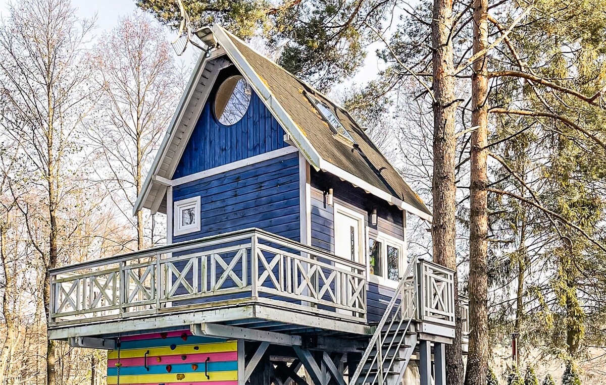 A charming two-story cottage is elevated on stilts amid tall trees. The structure features a blue exterior, wooden balcony railing, and large windows, allowing natural light. A colorful striped wall adds a playful touch, creating a welcoming atmosphere in the natural surroundings.