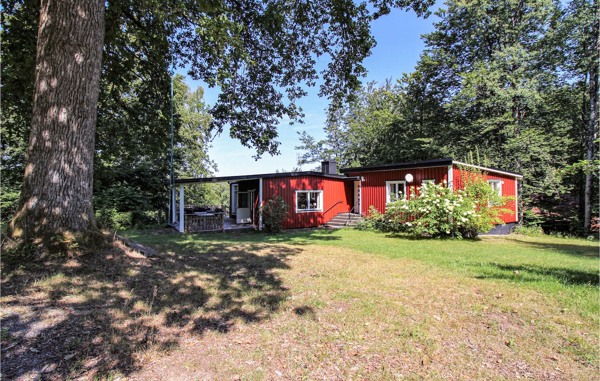 The exterior of a red cottage is surrounded by lush greenery and large trees. A spacious yard features a well-kept lawn, with a covered terrace visible on one side, providing shade and outdoor seating. The structure has a welcoming appearance, blending harmoniously with the natural landscape.