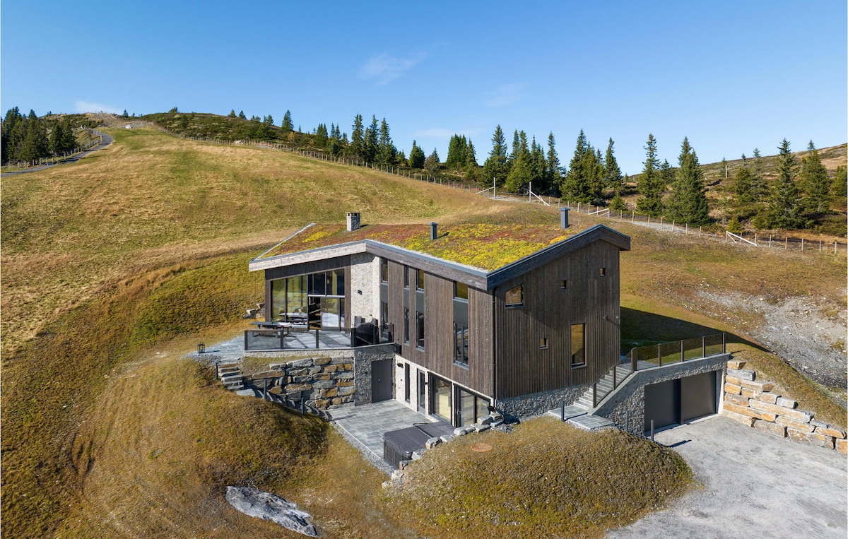 The modern holiday home features a wooden exterior with a green roof, harmoniously blending with the surrounding landscape. Large glass panels provide access to outdoor areas, while the elevated position offers panoramic views of the rolling hills and nearby trees.