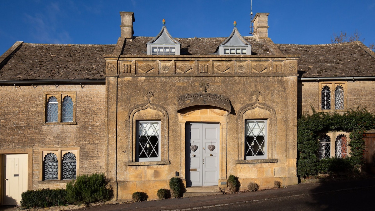 The exterior of Pineapple Spa Cottage features a stunning carved stone facade with intricate designs. The front door is framed by decorative elements, and unique windows with diamond-shaped panes add to its charm. The surrounding greenery complements the cottage's detailed architecture.