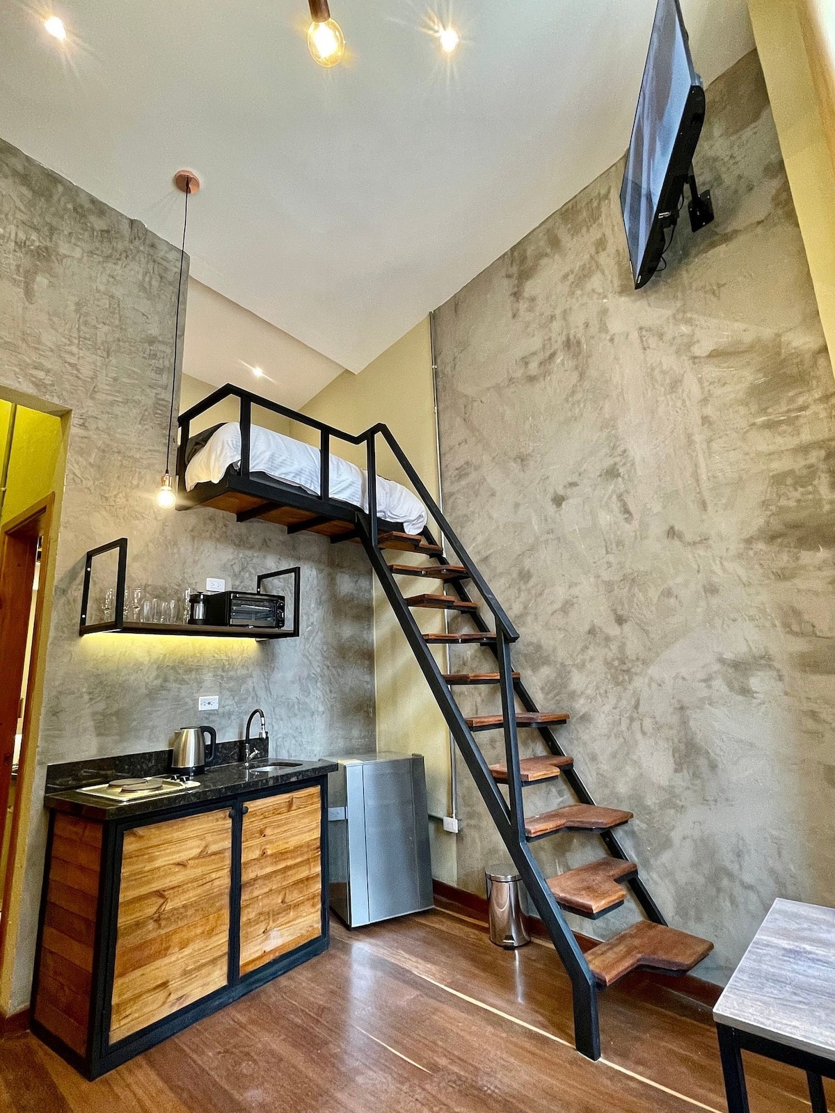 A modern studio apartment is showcased, featuring a compact kitchen area with wooden cabinets and a small refrigerator. A staircase leads to a lofted sleeping area, hinting at a cozy bed above. Natural light is aided by minimalist pendant lighting and a wall-mounted television.