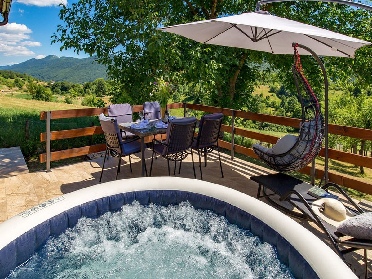 A private terrace showcases an outdoor dining area with a table and six chairs, accompanied by a shaded hanging chair. A hot tub is prominently featured, surrounded by greenery and offering views of the distant mountains under a clear blue sky.
