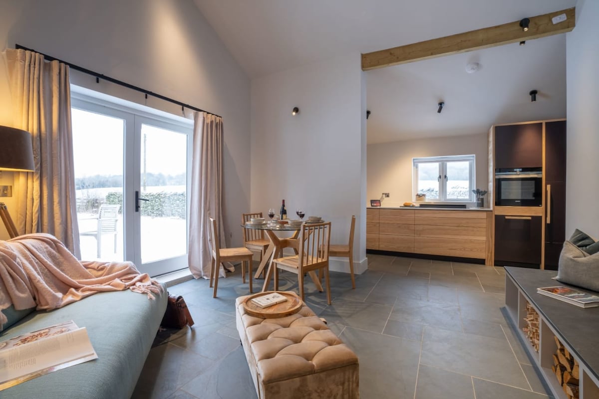 An open plan living space is displayed, featuring cozy seating with soft textiles and a dining area set for two. Light streams through glass doors that open to the patio, while a modern kitchen with wooden cabinetry is neatly arranged in the background.