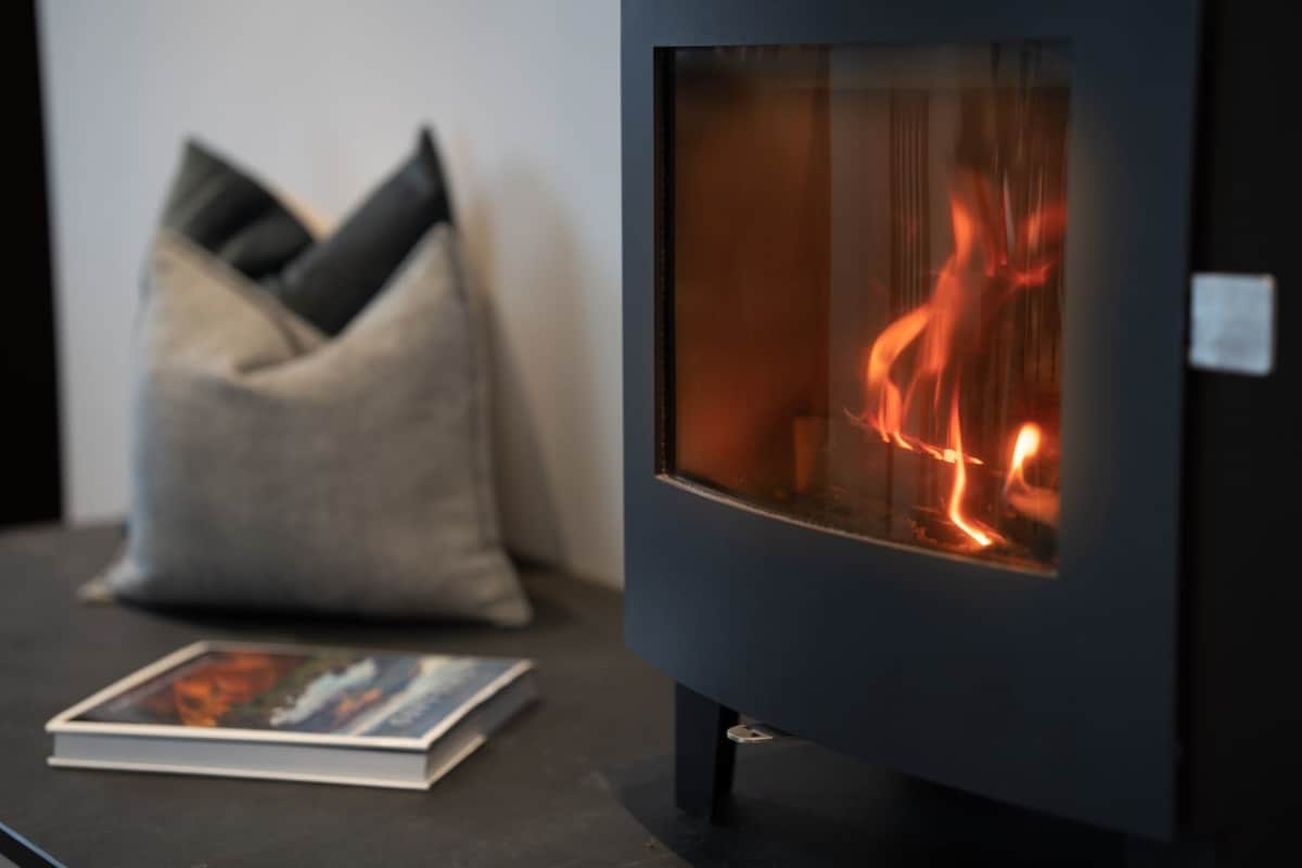 A cozy log burner is positioned beside a stack of soft cushions. An open book lies on a dark surface, creating an inviting atmosphere for relaxation. The warm glow of the flames adds a touch of comfort to the contemporary living space.