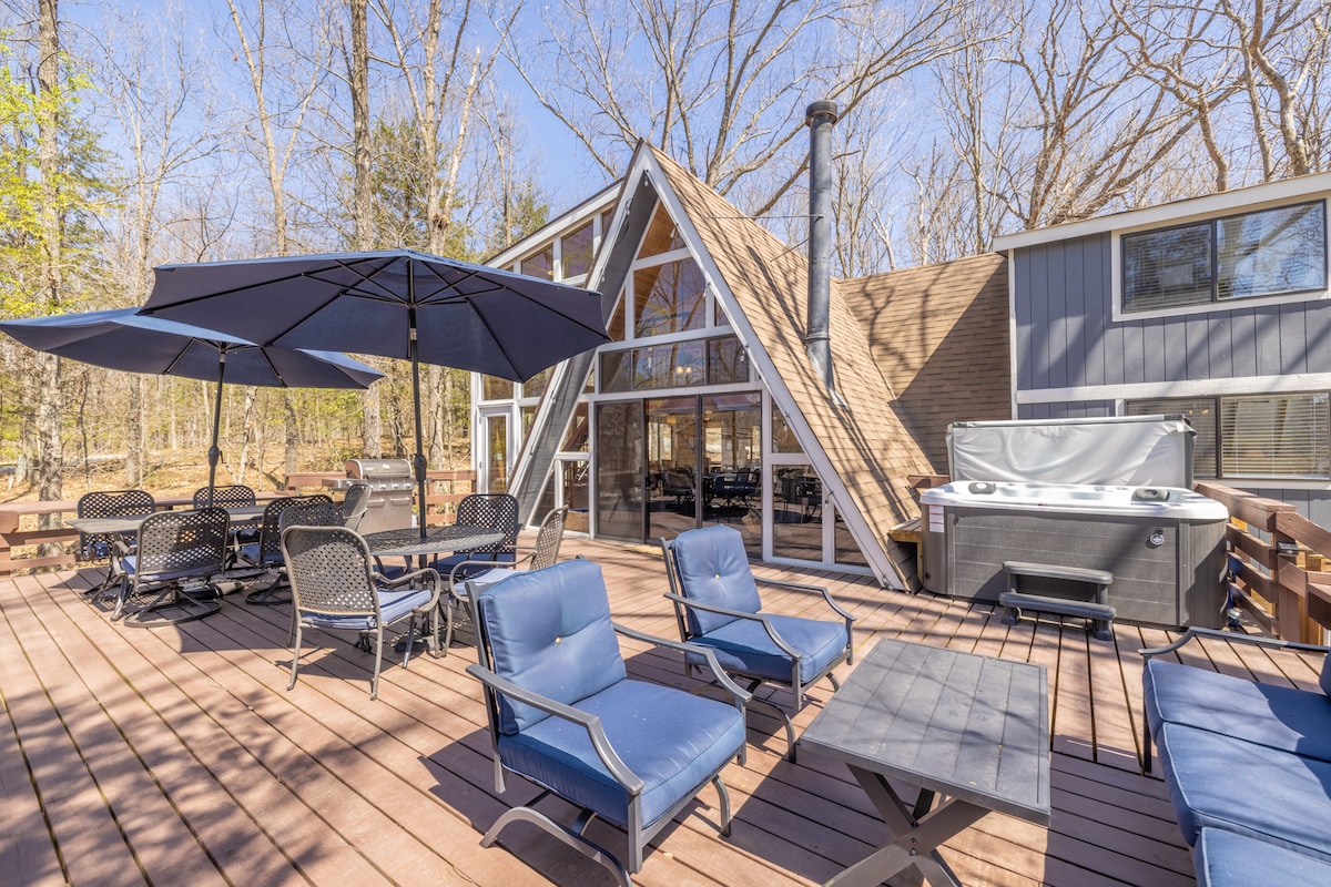 An expansive outdoor deck is featured, equipped with multiple seating areas, including dining tables and lounge chairs. A hot tub is positioned nearby, and large umbrellas provide shade. The chalet's distinctive a-frame structure and surrounding trees blend harmoniously with the outdoor space.