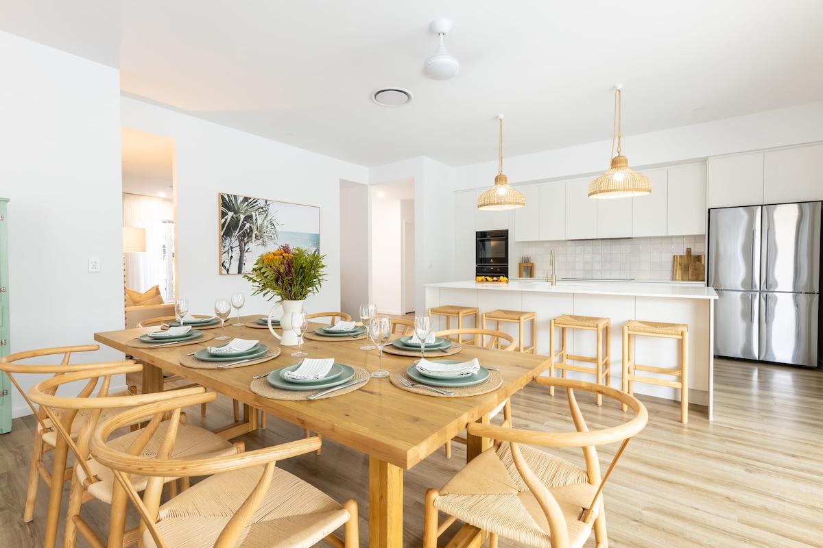 An open-plan living and dining area features a large wooden dining table set for eight, complemented by stylish woven chairs. Modern pendant lights hang from the ceiling, and a fully-equipped kitchen with sleek appliances is visible in the background.
