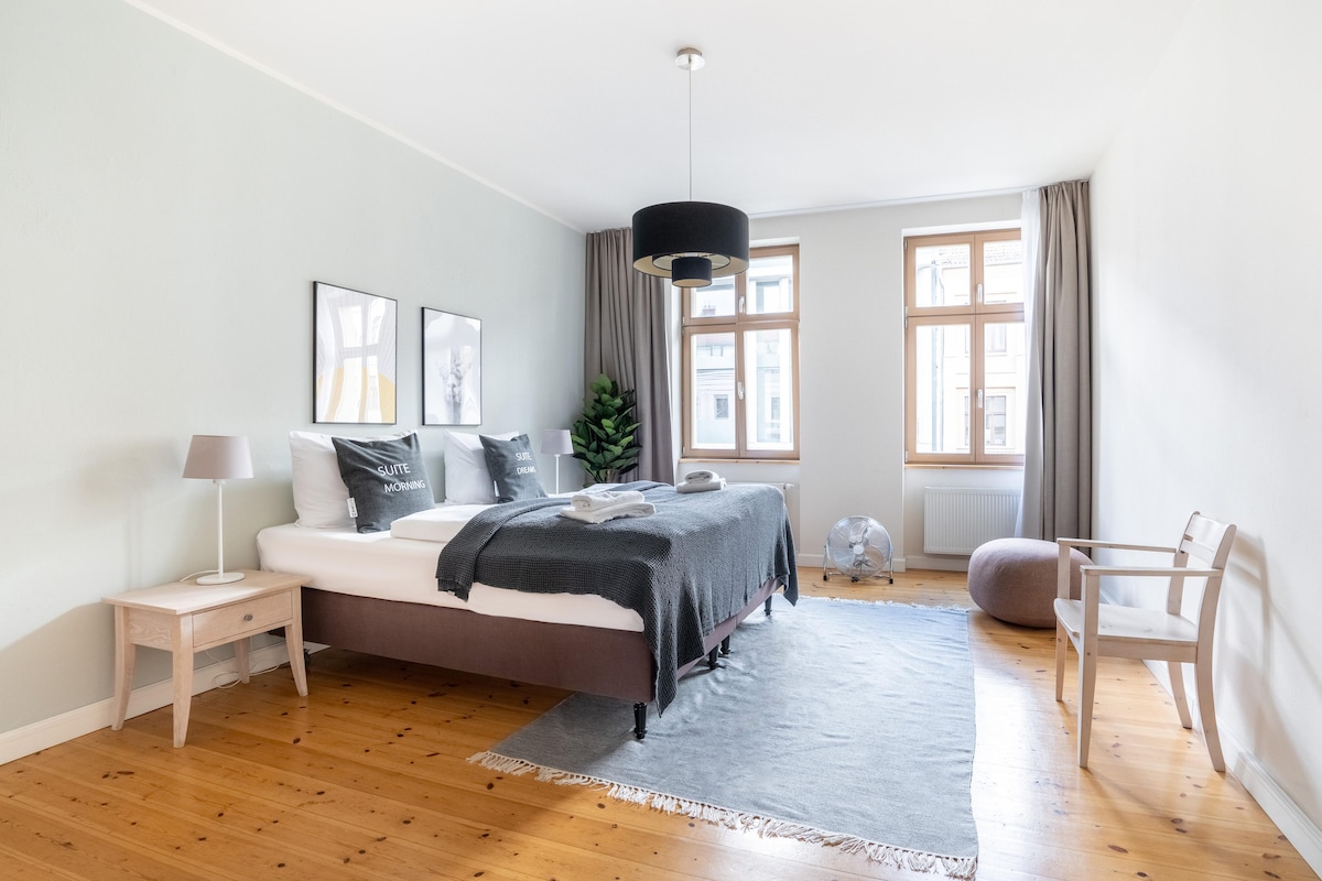 A spacious bedroom features a comfortable box-spring bed, adorned with soft blankets and pillows. Two large windows bring in natural light, and modern art pieces are displayed on the walls. A cozy armchair and a round pouf complement the wooden flooring and inviting decor.