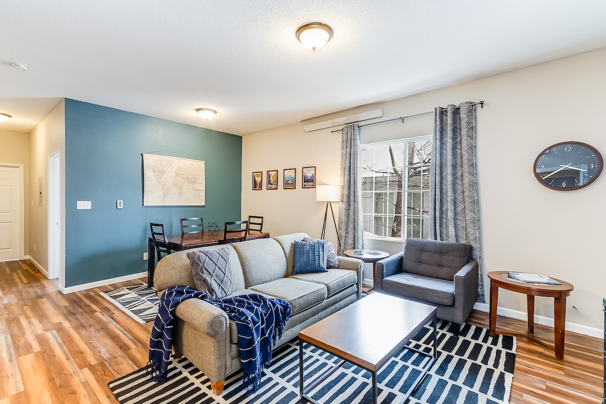 A spacious living room is presented with a comfortable grey sofa draped with a navy throw blanket, accompanied by a stylish armchair. A wooden coffee table sits center, with natural light flowing in from a large window framed by patterned curtains. A dining area with framed artwork is visible in the background.