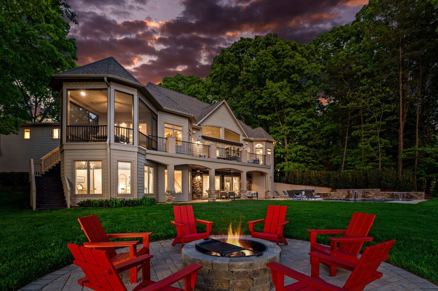 An expansive multi-level home is set against a twilight sky, featuring large glass windows that illuminate the interior. Red Adirondack chairs encircle a stone fire pit, inviting gathering. Lush greenery surrounds the property, and a heated pool is visible in the background.