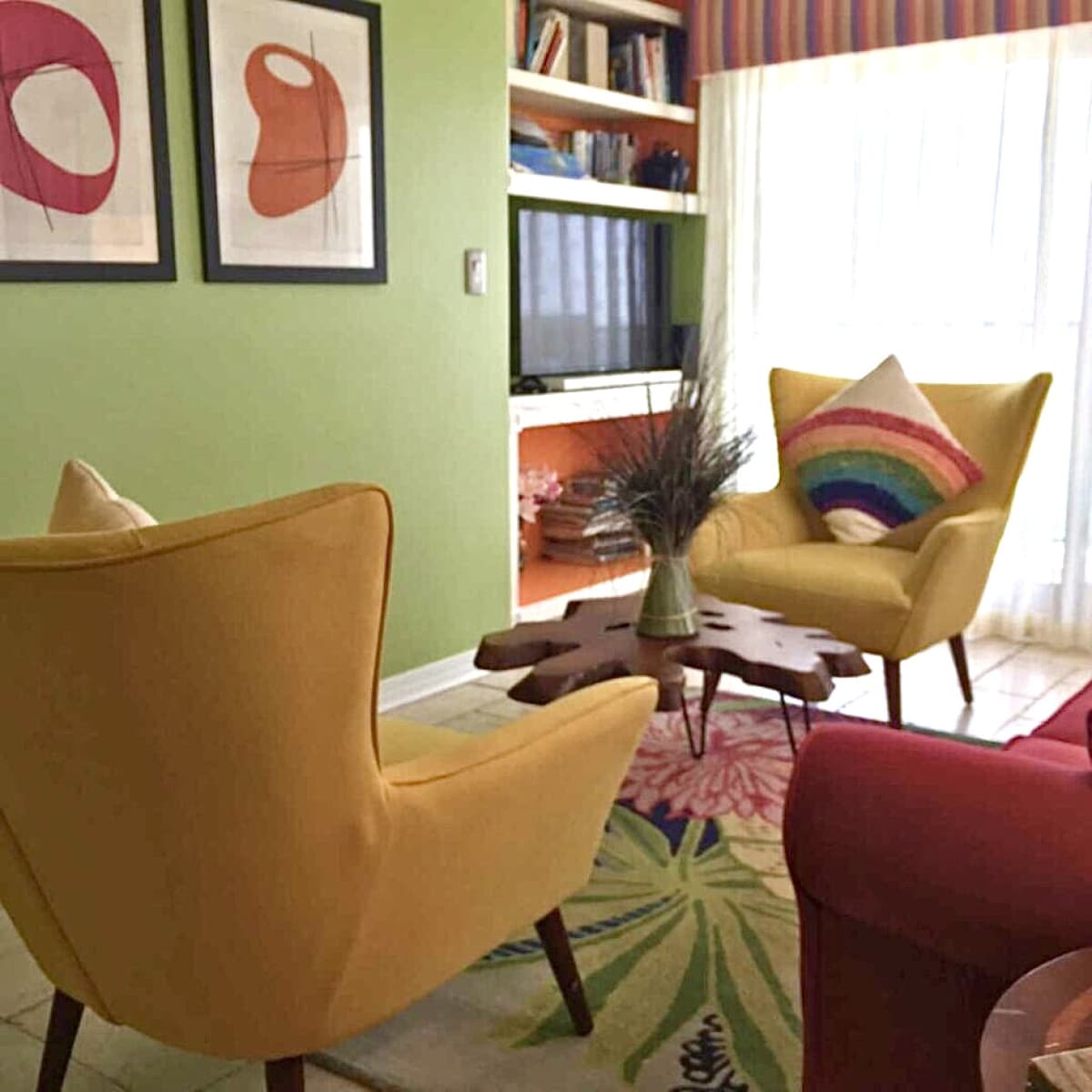 A vibrant sitting area features two yellow armchairs facing a colorful floral rug. A wooden accent table sits between them, adorned with decorative elements. Framed artwork hangs on green walls, while a bookshelf and television are visible in the background, complemented by sheer curtains allowing natural light.