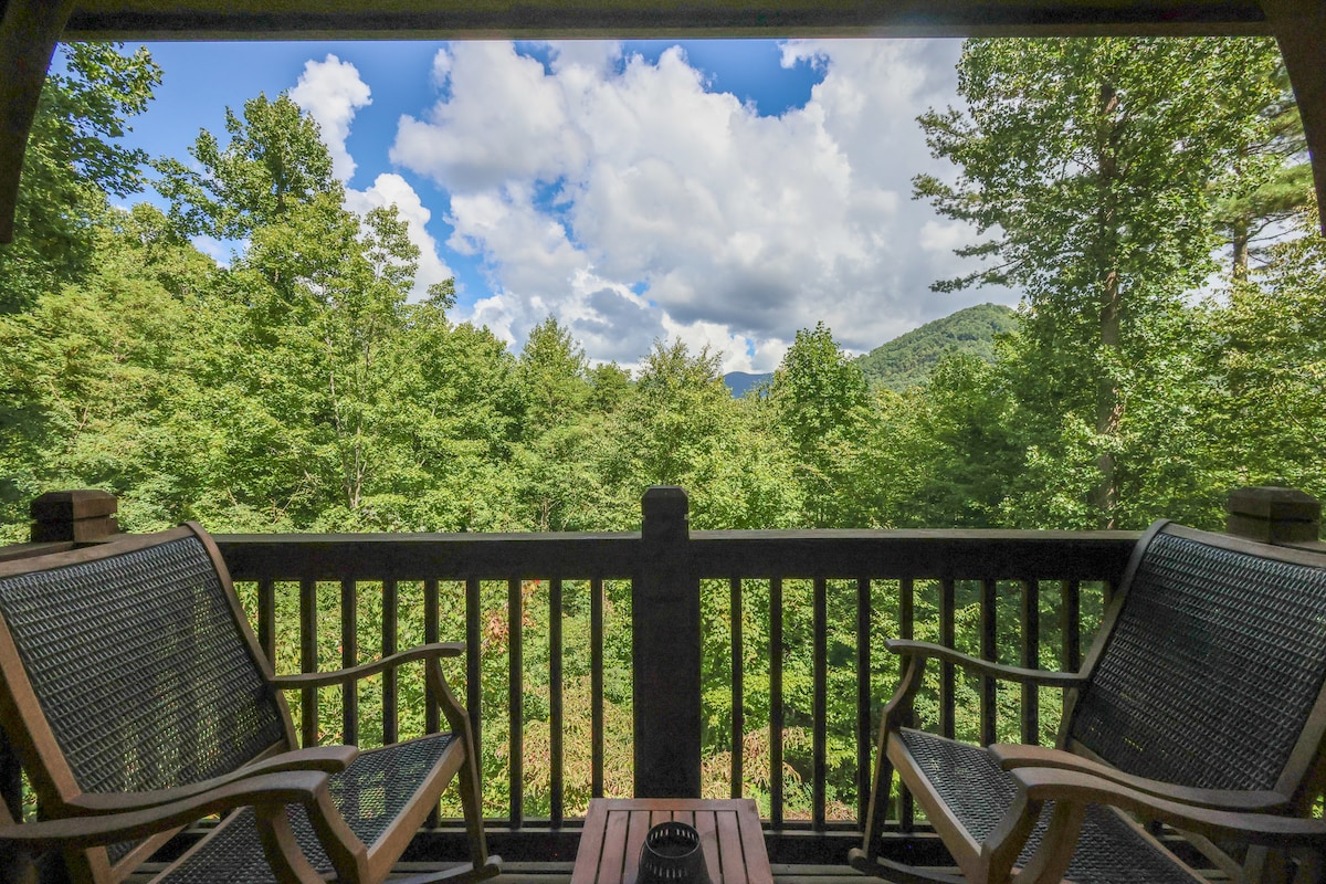 A private deck features two wooden chairs positioned on either side of a small table. Lush green trees extend into the background, with rolling mountains visible beneath a blue sky adorned with fluffy white clouds.
