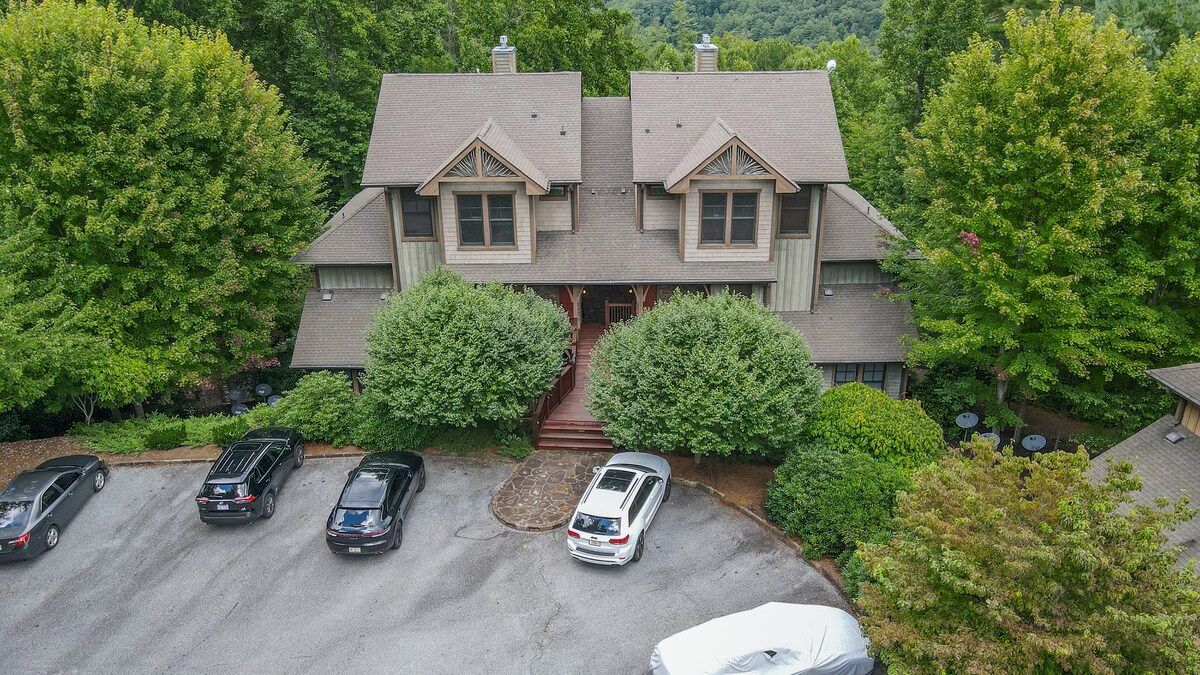 An aerial view of a craftsman-style upper-level condo is shown amidst lush greenery. Multiple parked vehicles are visible in a gravel lot, flanked by shrubbery and trees. The elegantly designed building features a prominent entrance with a staircase leading to the front door.