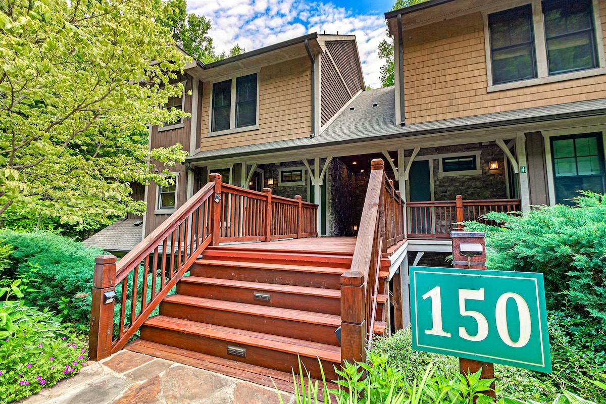 A welcoming entrance features a set of wooden stairs leading to a condo, framed by green foliage and a well-kept pathway. A sign displaying the number 150 is prominently placed near the lush landscaping, indicating the entryway.