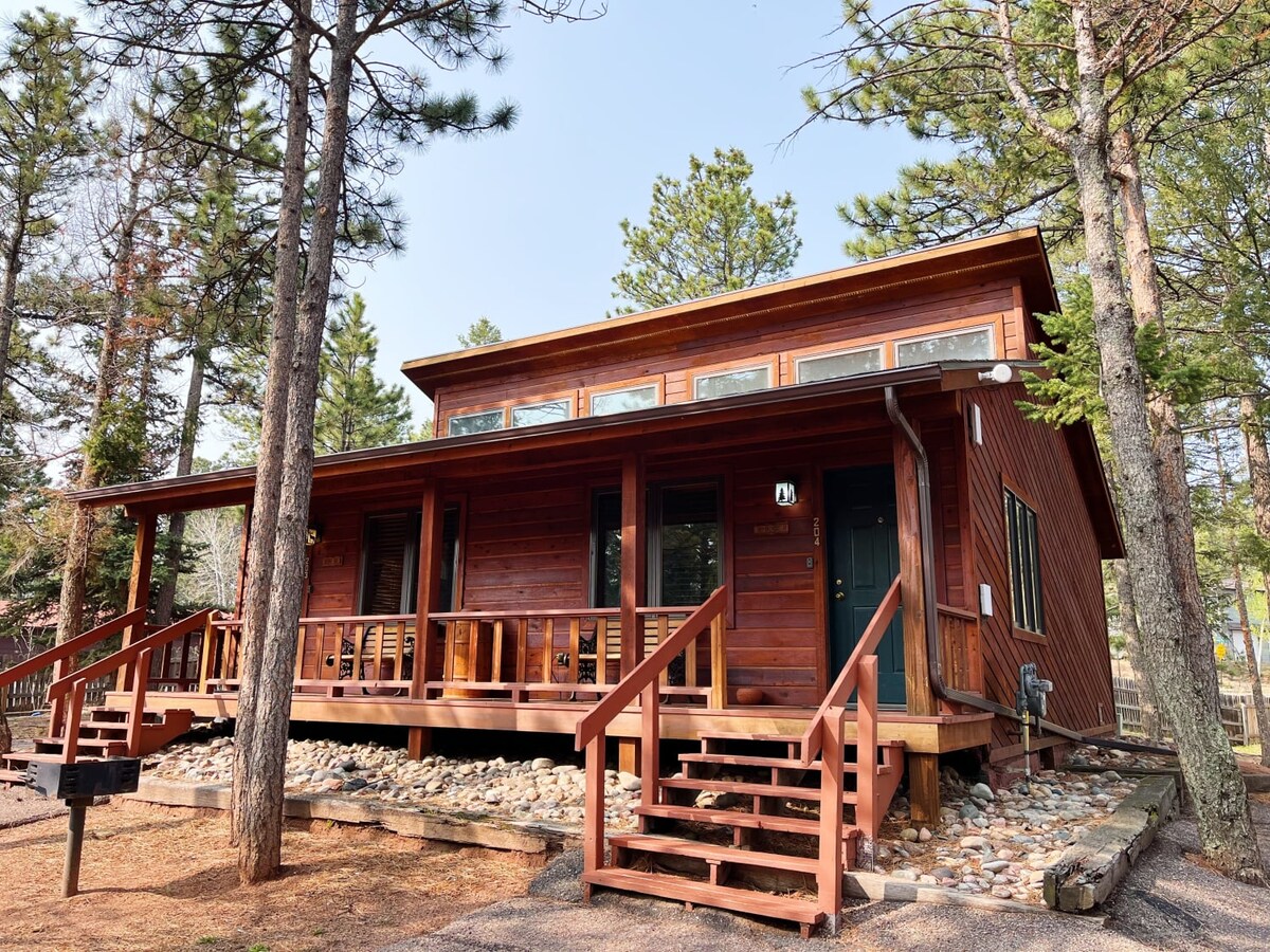 A rustic cabin structure is surrounded by tall pine trees, featuring a wooden porch with steps leading to the entrance. Large windows provide natural light, and a stone path enhances the outdoor appeal.