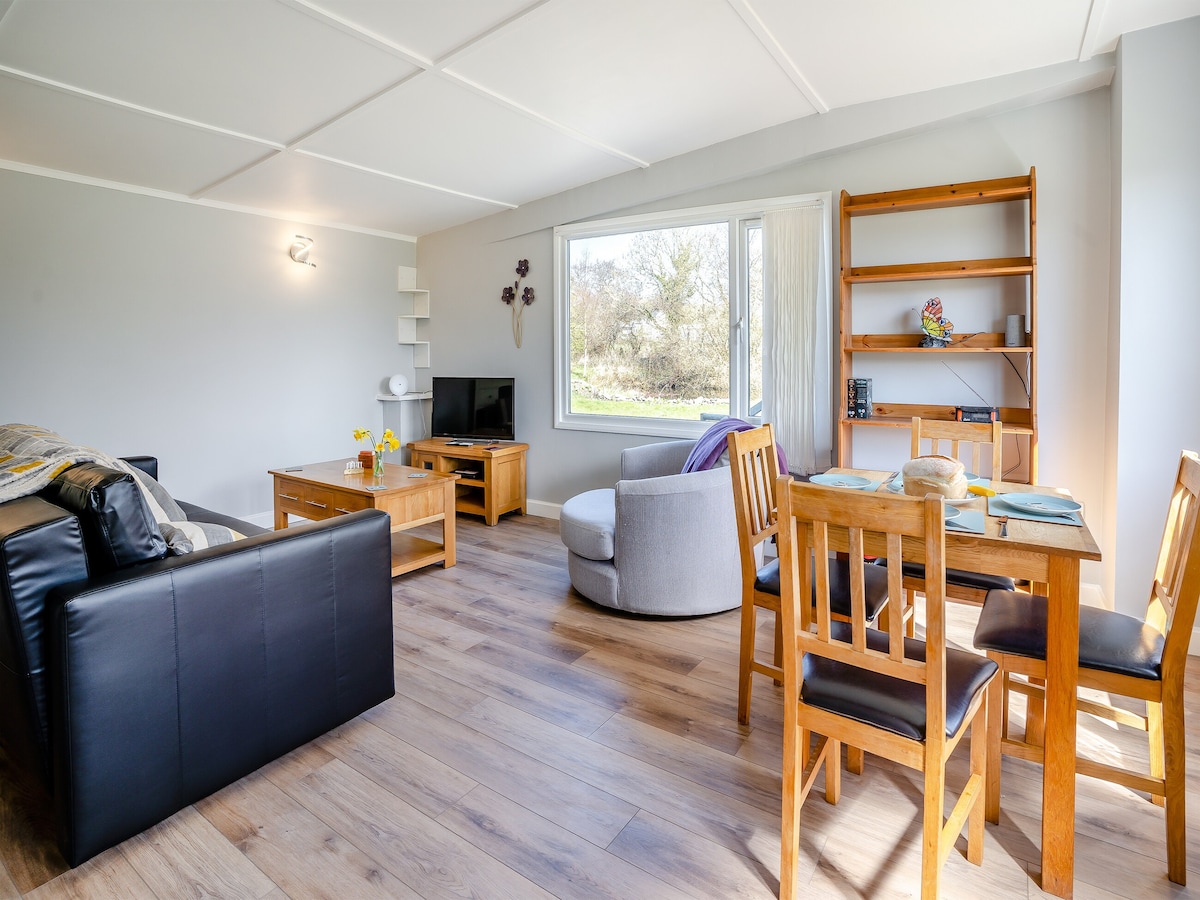 A spacious living and dining area features a comfortable sofa, an armchair, and a dining table set with chairs. A large window allows natural light to fill the space, providing views of the surrounding nature. A TV unit and shelving add functional charm to the room.