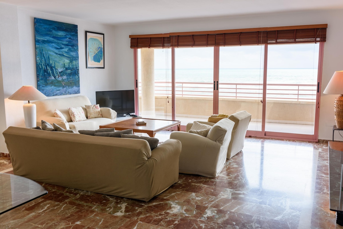 A spacious living area features two comfortable sofas, one of which serves as a bed. Large sliding glass doors open to a terrace with sea views, allowing natural light to brighten the space. A flat-screen TV and wooden coffee table complete the inviting setting.