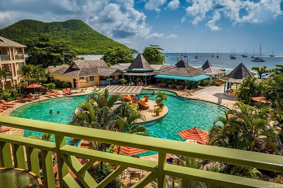 A scenic view reveals a large pool surrounded by lush tropical landscaping and lounge chairs. The pool area features umbrellas and structures, with the ocean and sailboats visible in the background, complemented by green hills under a partly cloudy sky.