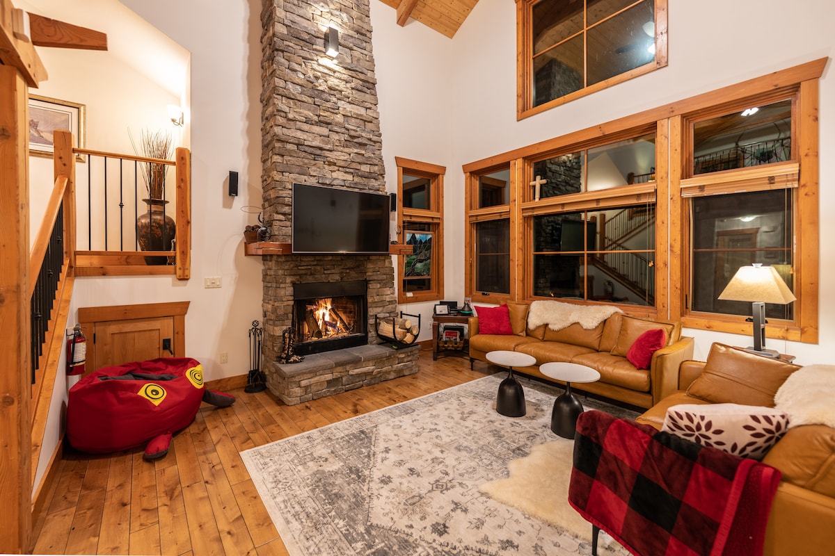 A spacious living area features a tall stone fireplace, providing warmth and charm. Large windows allow natural light to fill the room, while a comfortable beige sofa is complemented by two small black tables. A soft area rug lies underfoot, enhancing the inviting atmosphere.