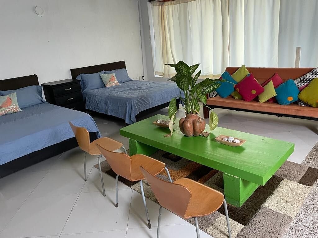 A spacious living area features two neatly made beds with blue linens and a colorful couch adorned with cushions. A green coffee table is centered among four wooden chairs, while natural light filters through large curtains, brightening the room.