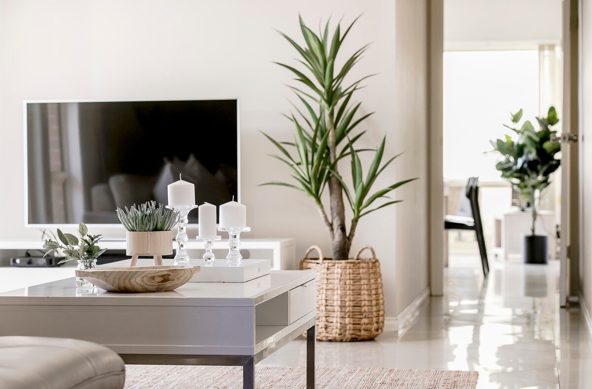 A contemporary living area features a sleek television positioned on a low table. A decorative arrangement of plants is visible, including a tall potted plant and smaller greenery. Light-colored walls enhance the airy feel, with natural light coming from a doorway leading to another room.
