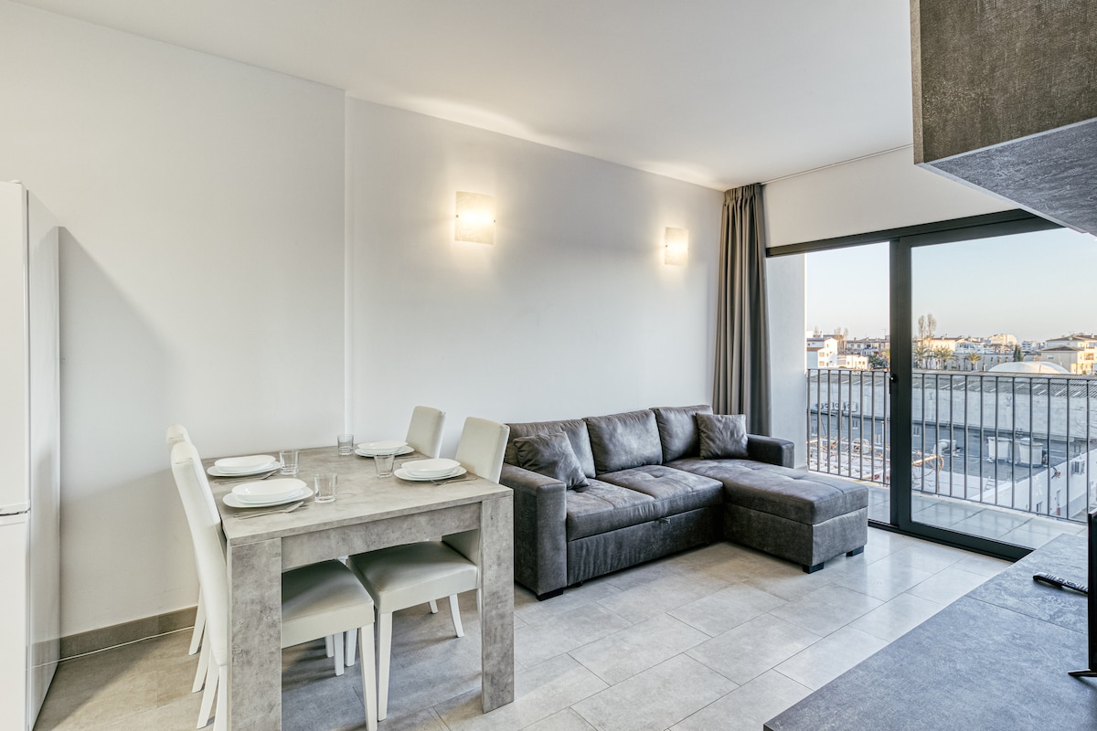 A modern living space is presented, featuring a spacious sofa and a dining table with four chairs. Large windows allow natural light to fill the room, while neutral tones create a calm environment. The balcony is accessible, offering views of the surroundings.