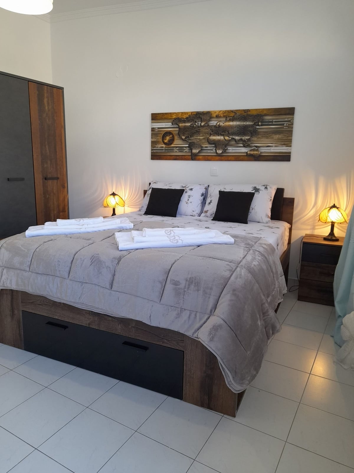 A cozy sleeping area is featured with a king-size bed, adorned with soft linens and neatly arranged towels. Warm light from bedside lamps illuminates the room. A modern painting hangs above the bed, and a wardrobe is positioned nearby for storage.