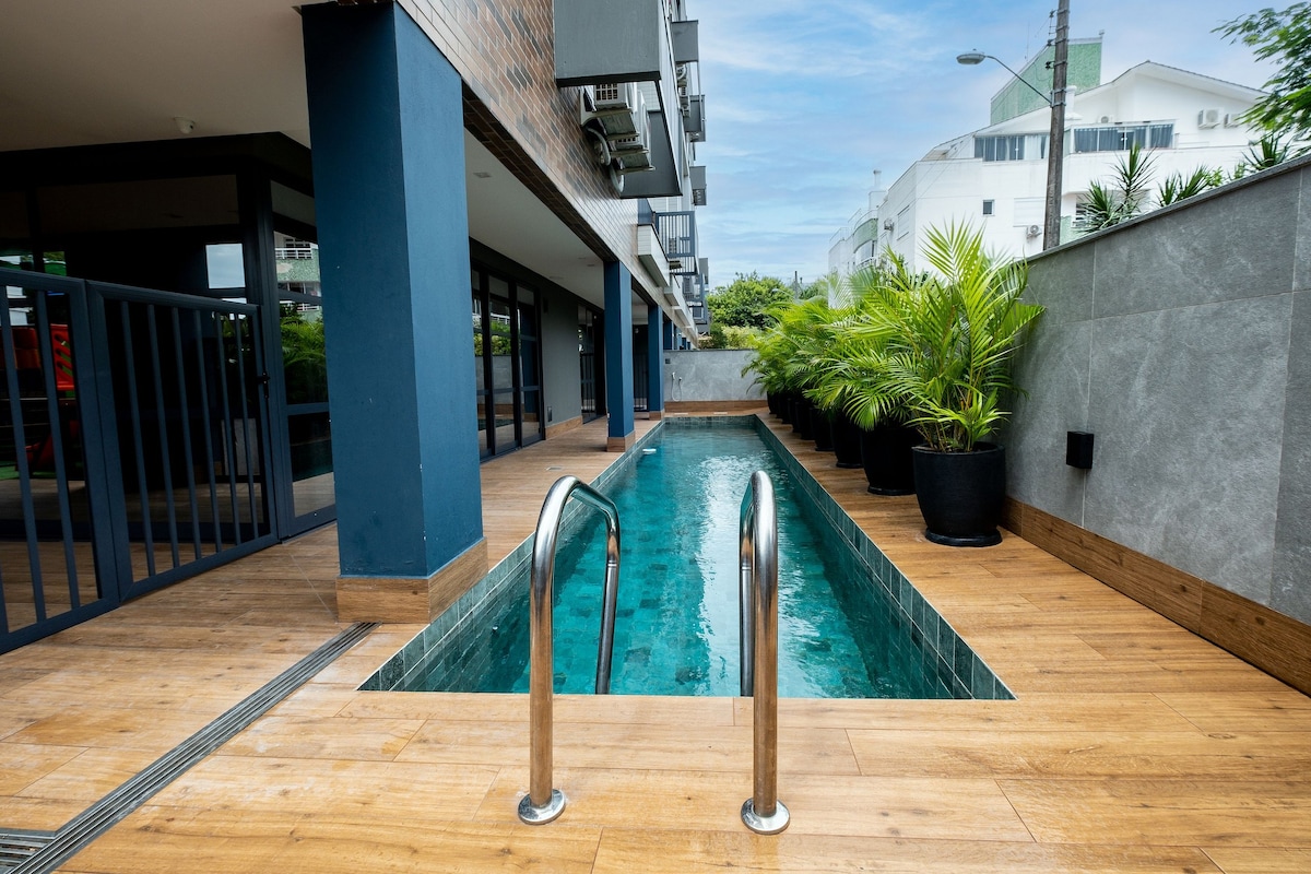 A narrow swimming pool is presented alongside a wooden deck. Rows of tall green plants are positioned in black pots against the gray walls, offering a touch of nature. The area is framed by the modern buildings of the property, with ample natural light highlighted.