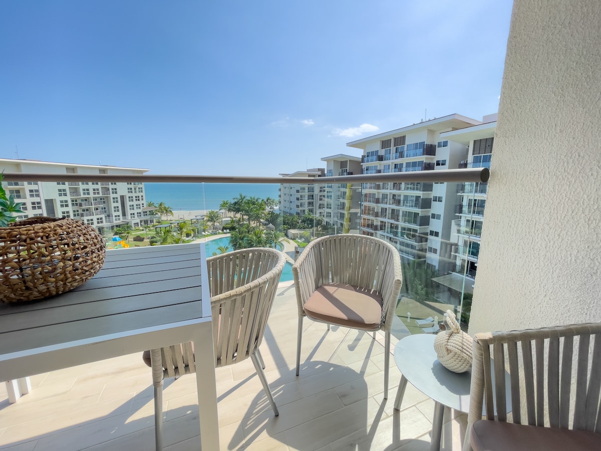 A private balcony offers a view of the ocean and surrounding complex. Two round chairs and a small table are arranged comfortably. Lush greenery and a swimming pool are visible below, contributing to the serene atmosphere of the location.