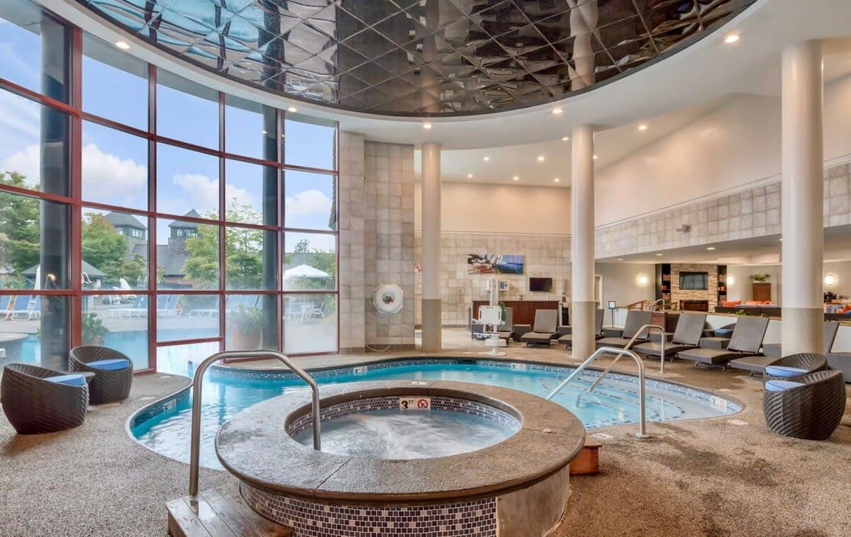 An indoor hot tub is surrounded by a spacious area with lounge chairs and large windows that allow natural light to fill the space. The ceiling features a geometric design, and the soothing water is visible within the rounded tub.