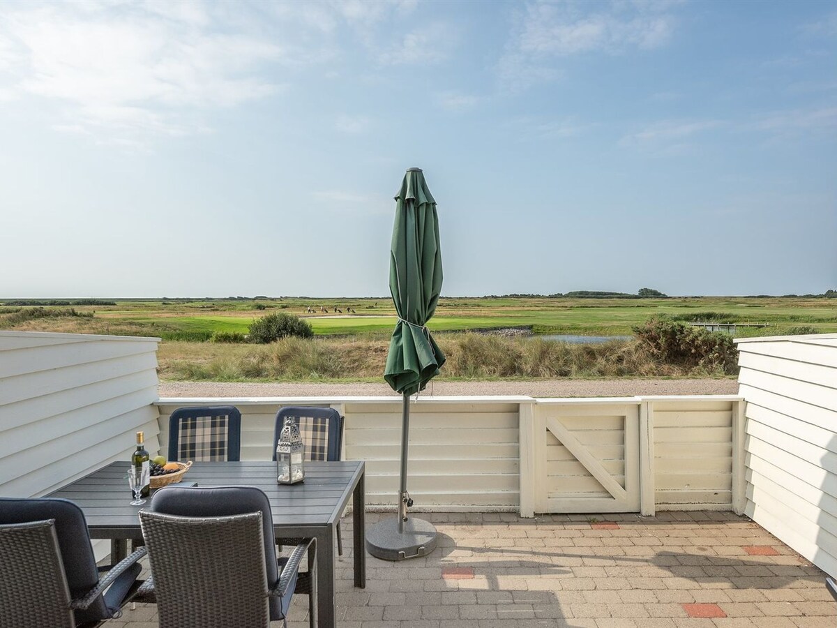 A covered outdoor dining area features a table and four chairs, overlooking a natural landscape. A green umbrella is positioned beside the table, providing shade. The serene view includes open fields and distant greenery under a clear sky.