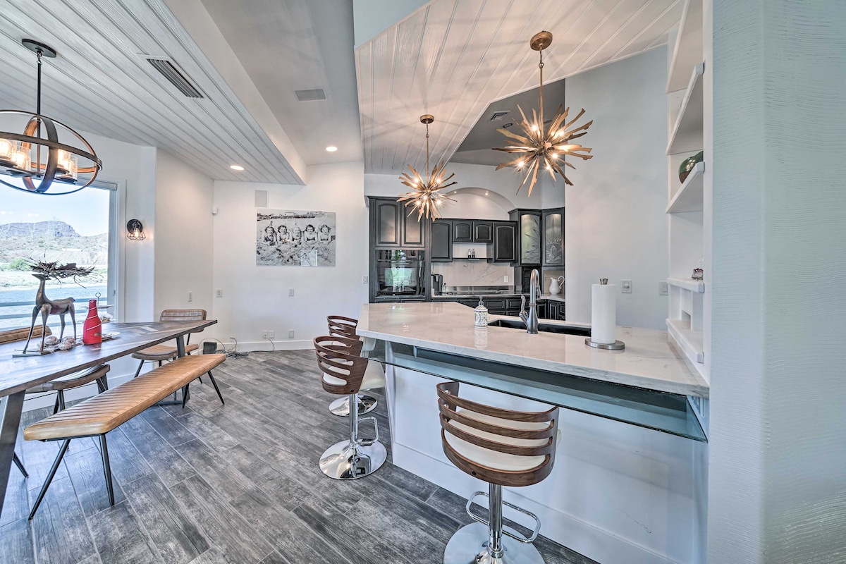An open-concept kitchen and dining area features a large dining table made of wood alongside sleek barstools at a kitchen island. A contemporary light fixture with starburst design hangs above the island, illuminating neutral tones and ample natural light from nearby windows.