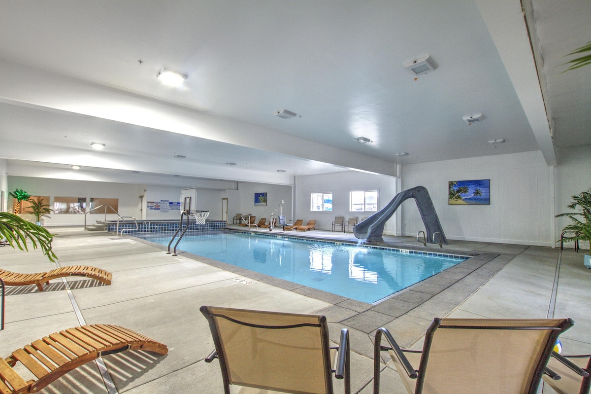 An indoor recreation area features a swimming pool with a slide, surrounded by lounge chairs. Bright overhead lights illuminate the space, and large windows allow natural light to enter. Artwork adorns the walls, enhancing the inviting atmosphere.