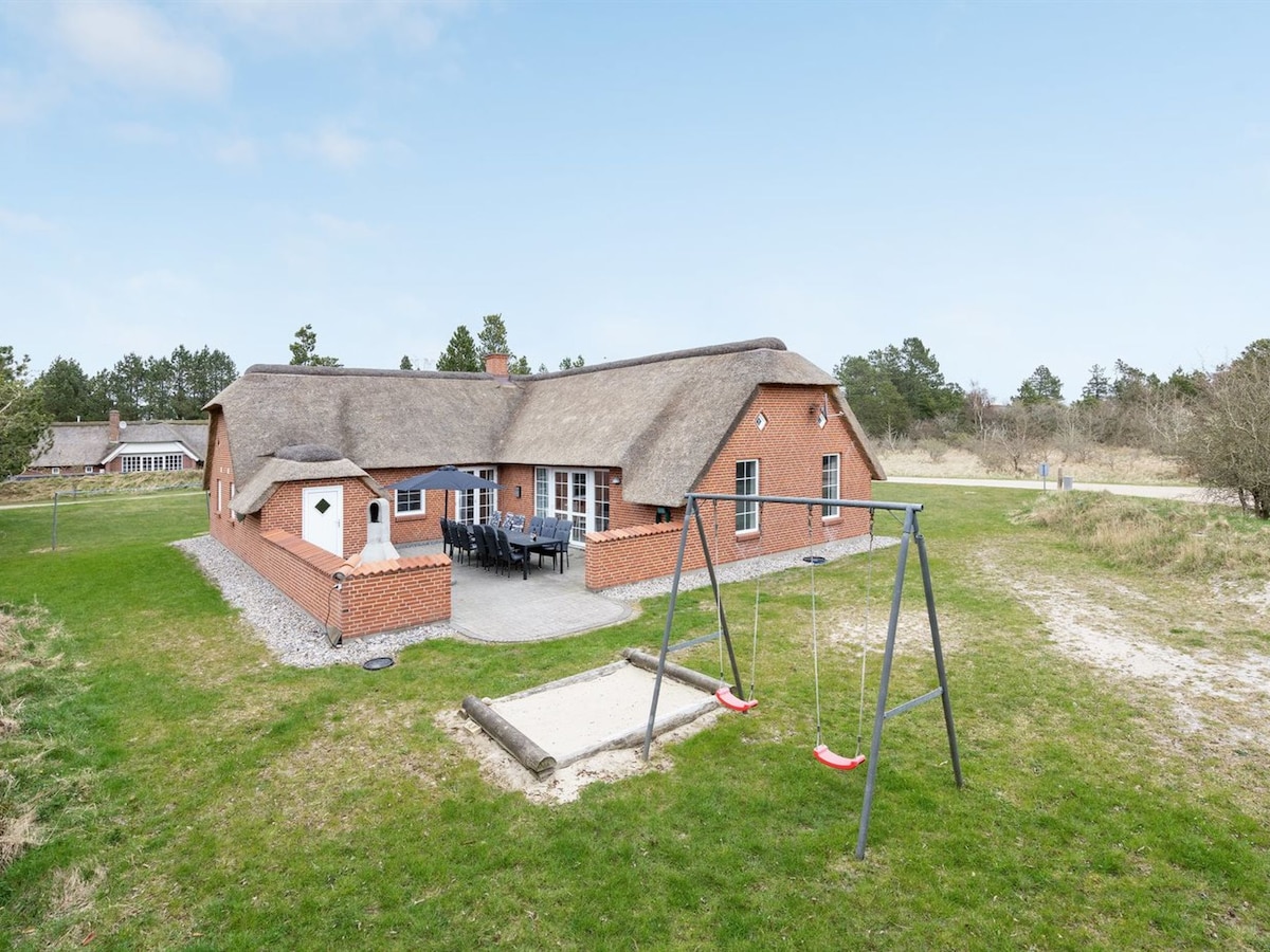 The exterior of the house features a traditional thatched roof surrounded by greenery. A patio area is visible with outdoor seating for dining. A children's playground area includes a swing set and a sandpit, offering space for outdoor leisure.