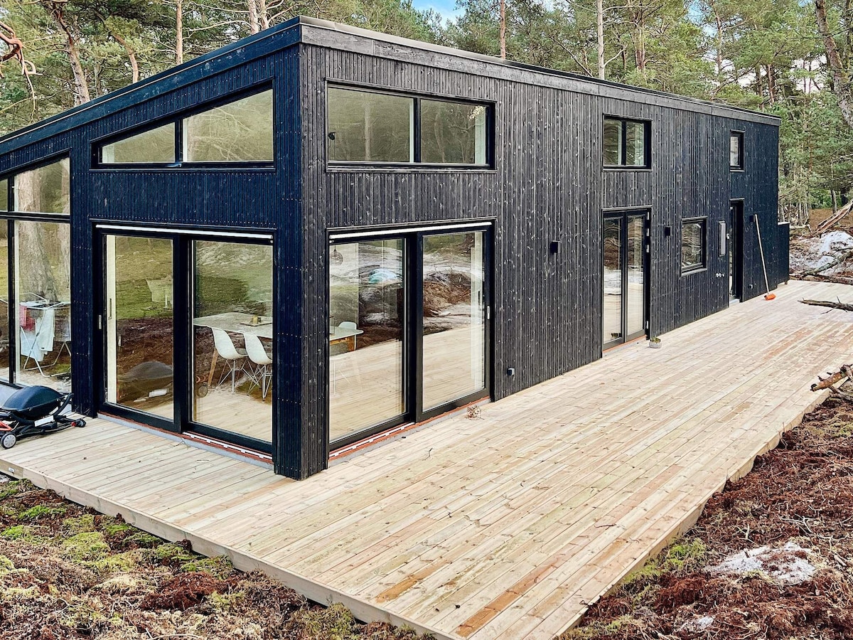 A modern holiday home is set among trees, featuring large floor-to-ceiling windows that provide ample natural light. A spacious wooden terrace extends outward, creating an inviting area for outdoor relaxation. The sleek black exterior contrasts with the natural surroundings.