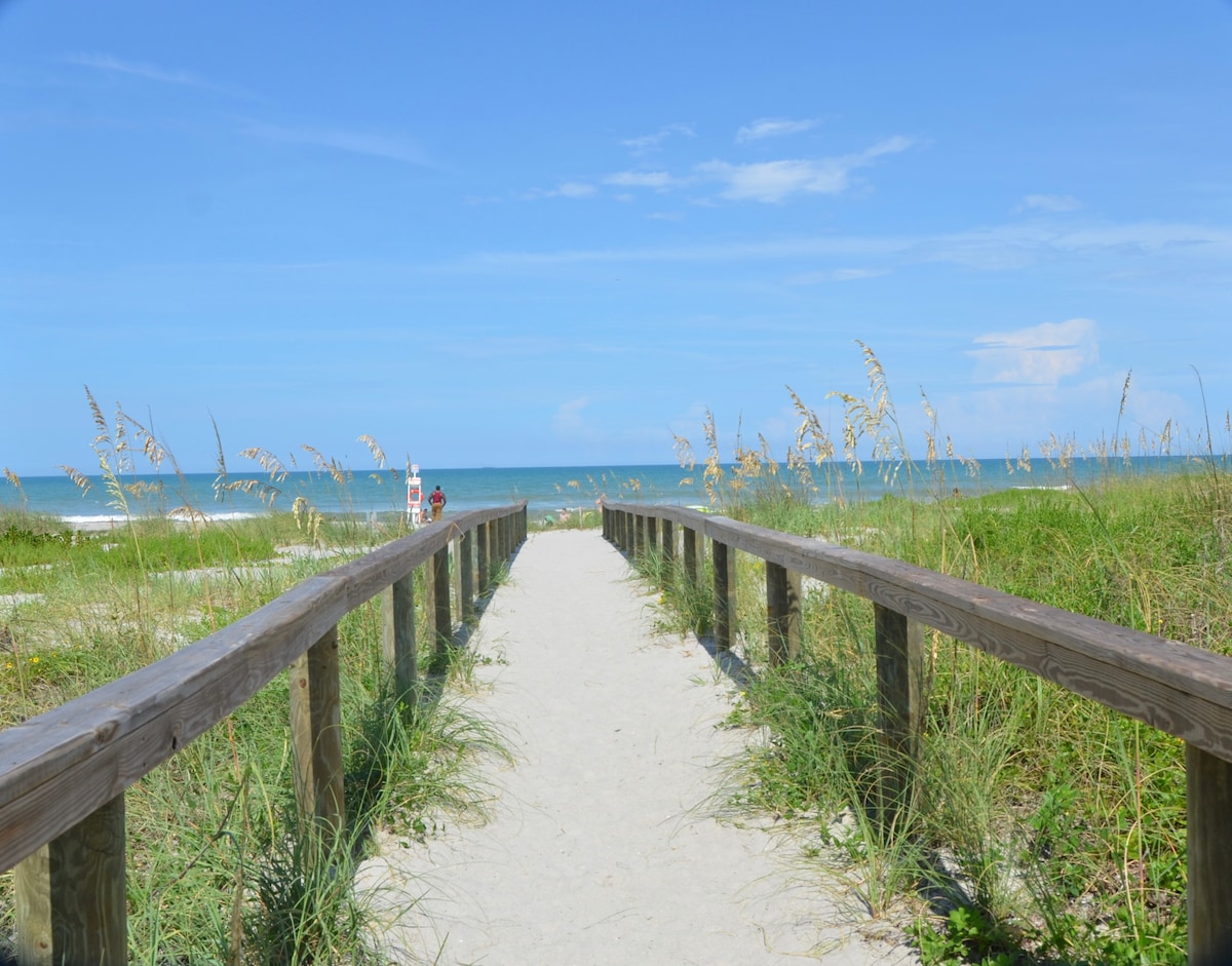 A wooden boardwalk leads through sandy terrain, flanked by tall grass, to the ocean. The serene blue waters stretch into the horizon under a clear sky, creating an inviting pathway to the beach.