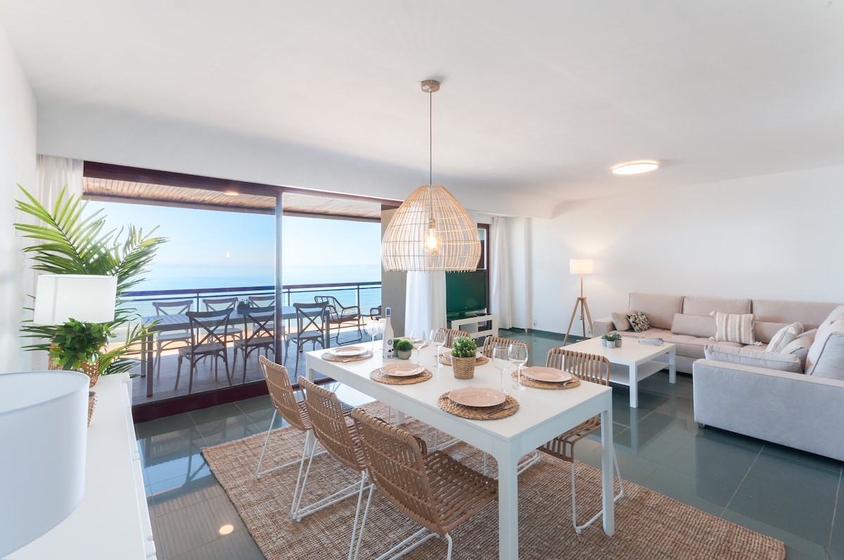 A light and airy living space features a white dining table set for six, surrounded by wicker chairs. A plush sofa sits nearby, with large windows offering expansive views of the sea. Natural light floods the room, enhancing the modern decor.