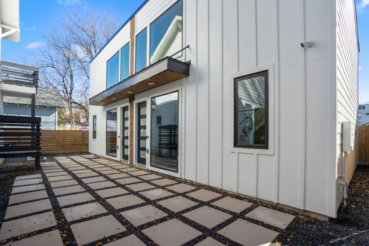An exterior view highlights a modern building featuring large glass doors and two smaller windows. A paved area with square stones is visible, bordered by gravel and foliage. The setting appears well-maintained, with a mix of natural light and shadows across the surface.