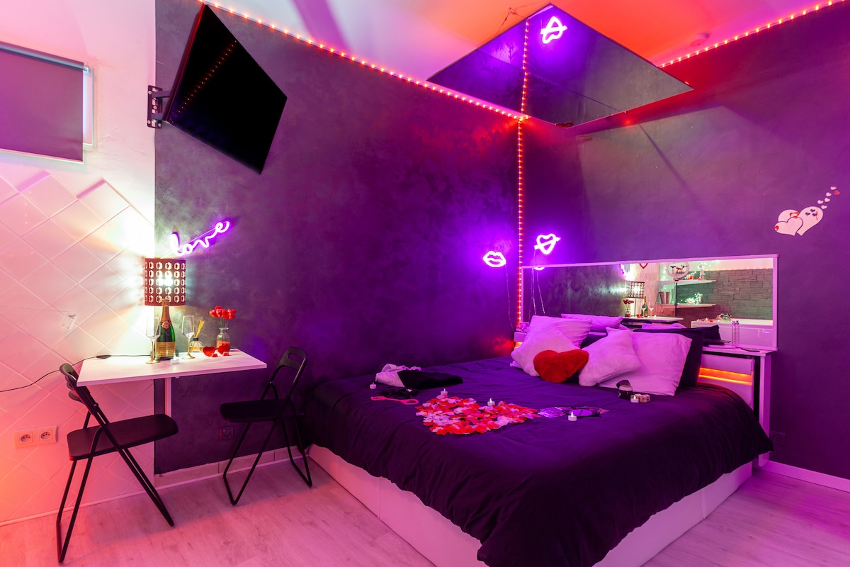 A stylish studio room features a spacious bed adorned with decorative pillows and rose petals. A small table with two chairs is positioned nearby, and LED lighting adds a soft glow to the walls. A television and mirrored accents enhance the modern ambiance of the space.