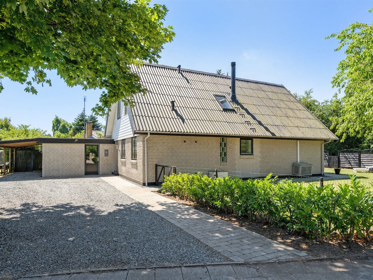 The two-story house is surrounded by a well-maintained garden and features a gravel parking area. Notable elements include a steeply pitched roof, multiple windows, and a covered entryway, providing a welcoming appearance in a serene outdoor setting.