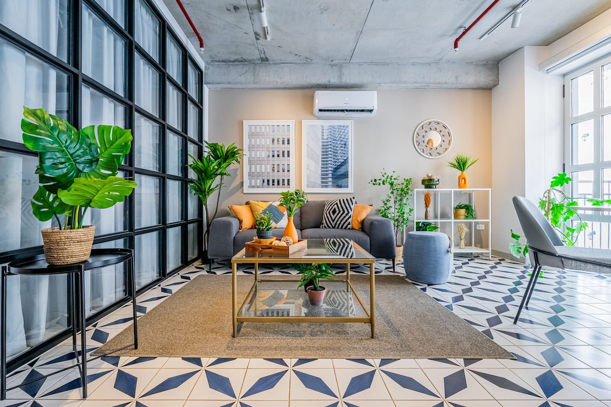 A bright living area is showcased with modern furnishings on a patterned tile floor. A glass coffee table sits centrally, surrounded by a cozy sofa, accent chairs, and decorative plants. Natural light fills the space through large windows, highlighting the contemporary decor.