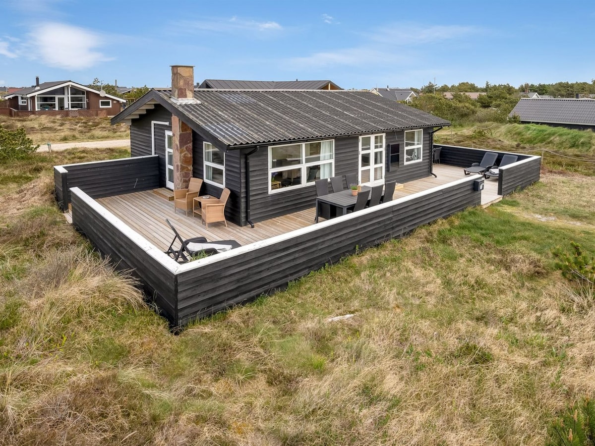 A single-family house is surrounded by a grassy landscape, featuring a spacious wooden terrace. The exterior is clad in dark wood, with large windows allowing natural light inside. Outdoor seating areas are arranged on the terrace, offering space for relaxation.