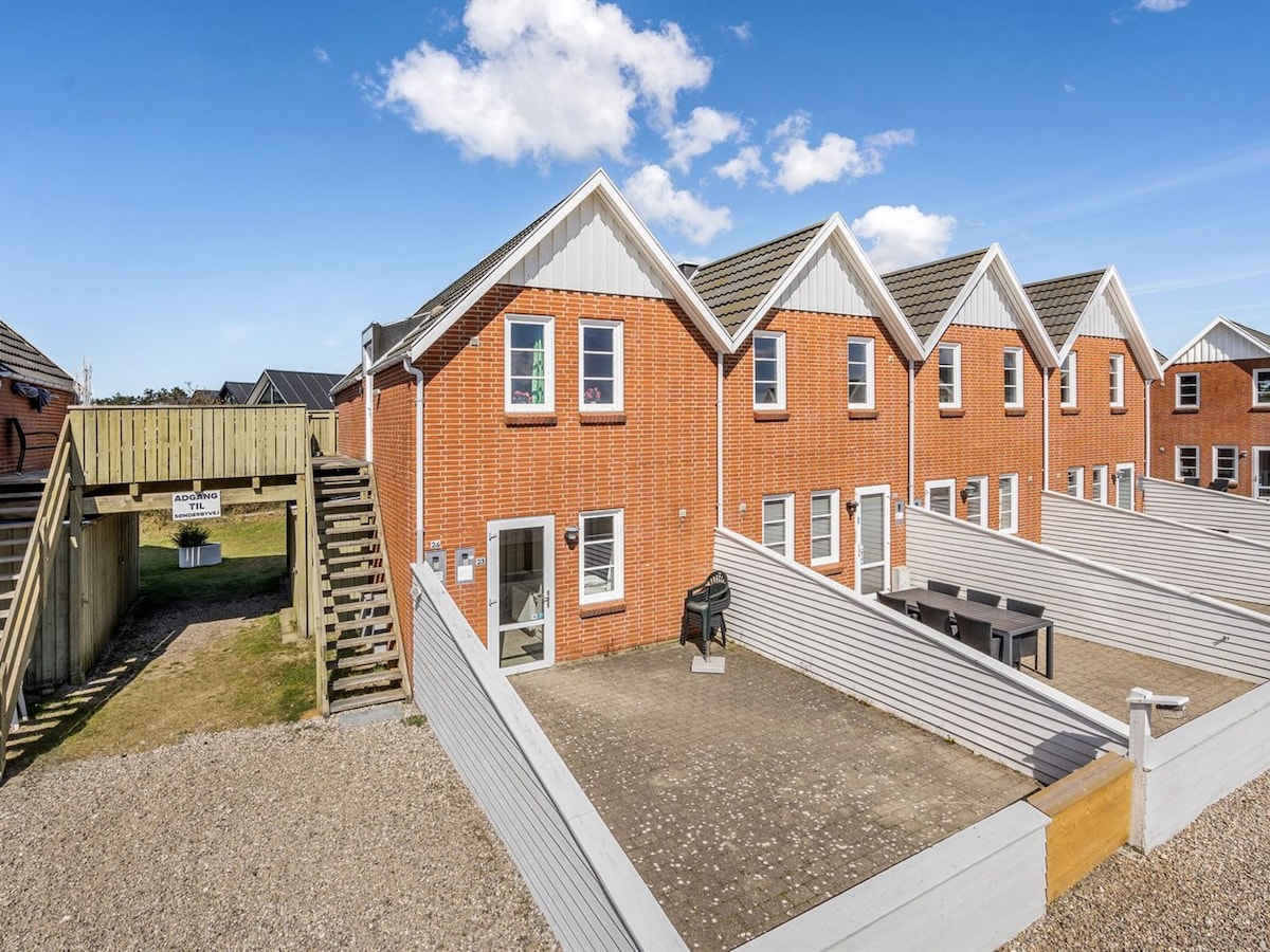 A row of two-story brick apartments is displayed, each featuring multiple windows. A pathway leads to a staircase, providing access to elevated outdoor areas. Outdoor furniture is visible, with a barbecue unit positioned to the side, while an expansive lawn area is behind.