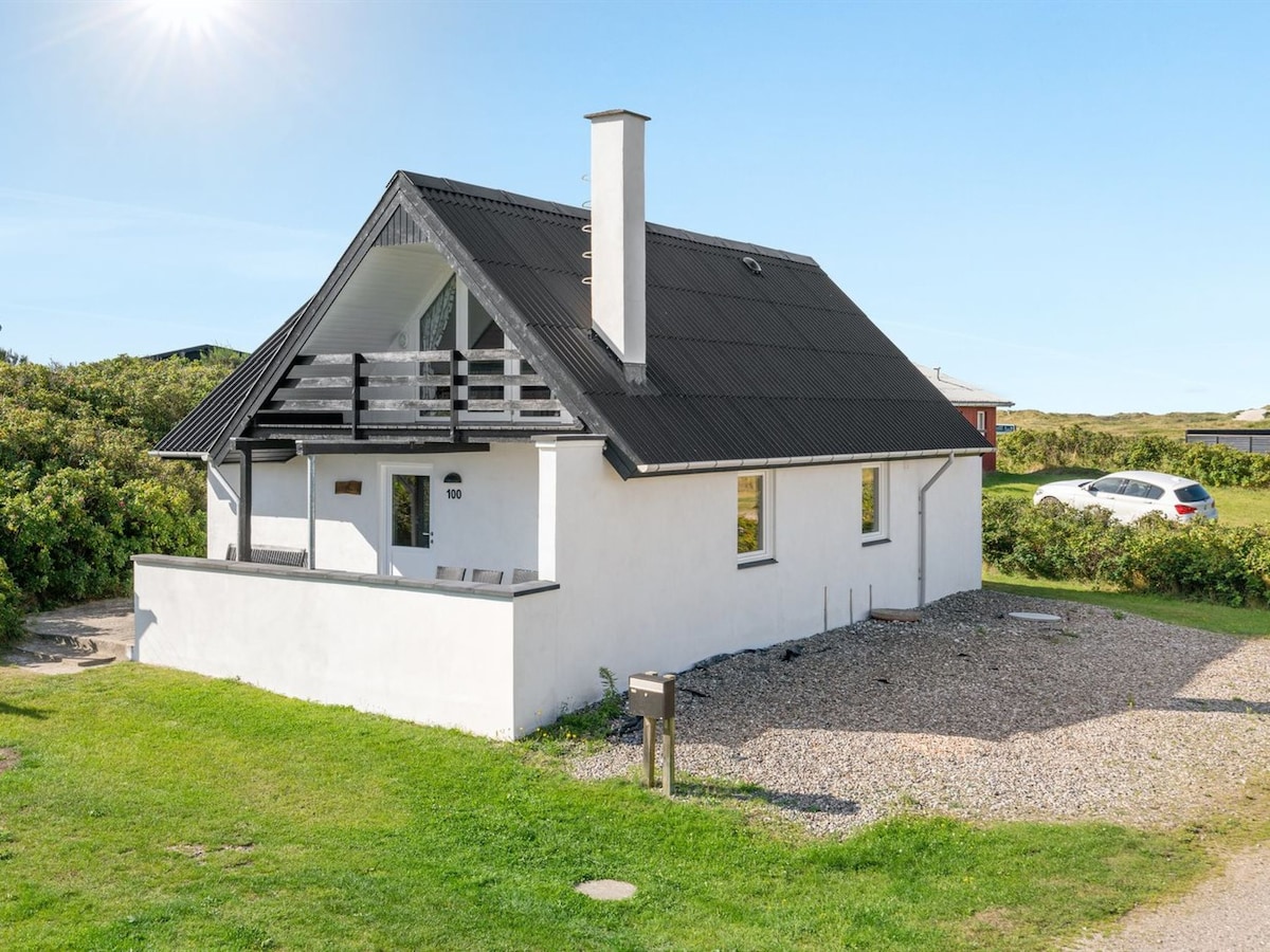 The exterior of the house is displayed, featuring a white facade and a black roof. A chimney is visible on the roof. A gravel area surrounds the house, with green grass and natural vegetation nearby. The parking area can be seen in the background.