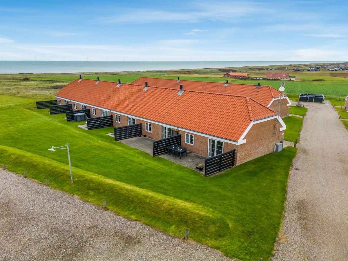 A spacious two-story building with a red-tiled roof is set on a green lawn, surrounded by open fields. The property features outdoor seating areas with tables and chairs. The nearby path leads to a gravel drive, contributing to the serene environment adjacent to the sea.